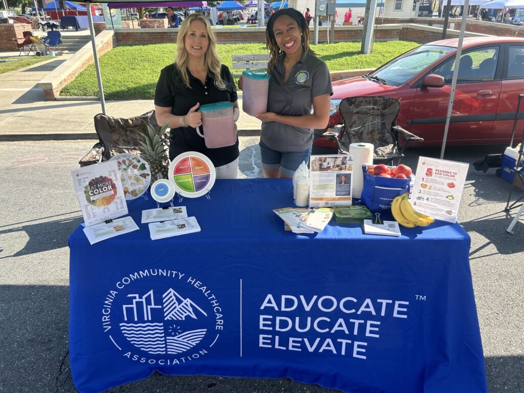 Angela and Adrienne hold up pitchers of smoothies behind a table covered in nutrition information and healthy snacks.