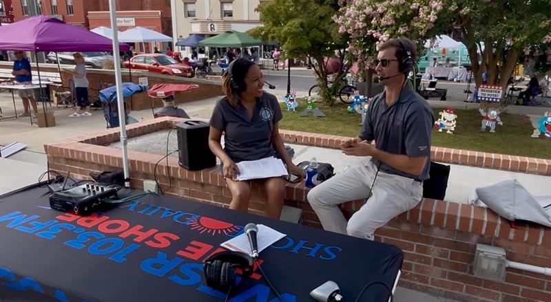 Adrienne speaks with a radio host in a town square, both are wearing radio headsets.