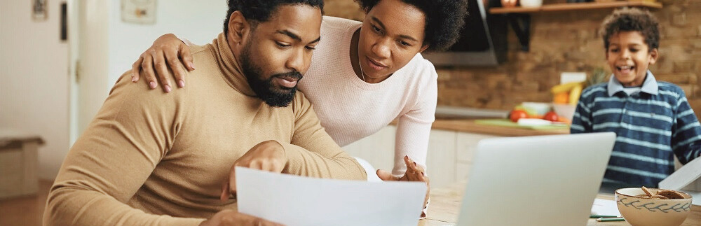 A Black man seated at a table in front of a laptop looks at a paper; A Black woman leans over his shoulder.