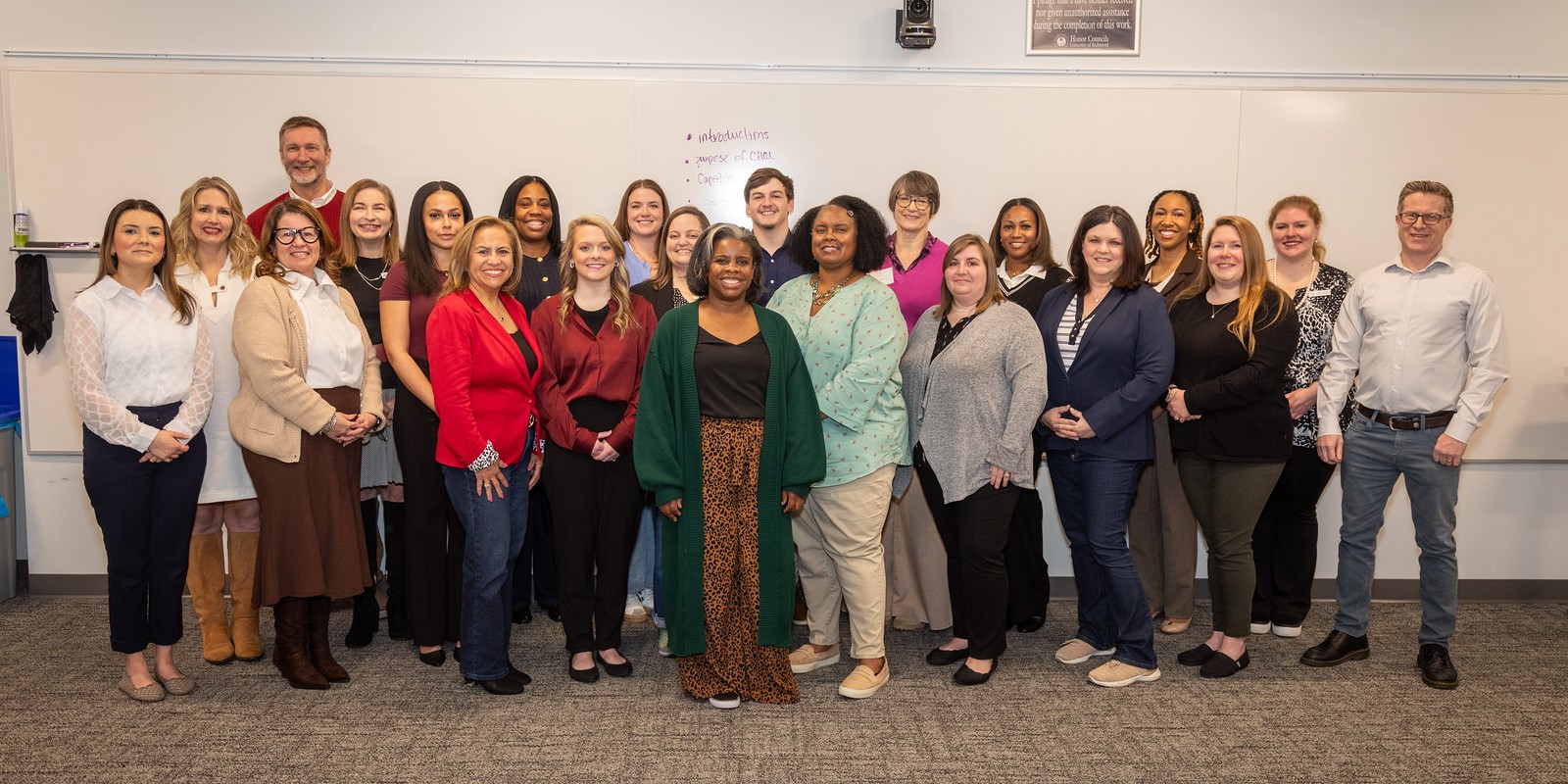 A group of about 25 smiling people poses in front of a whiteboard.