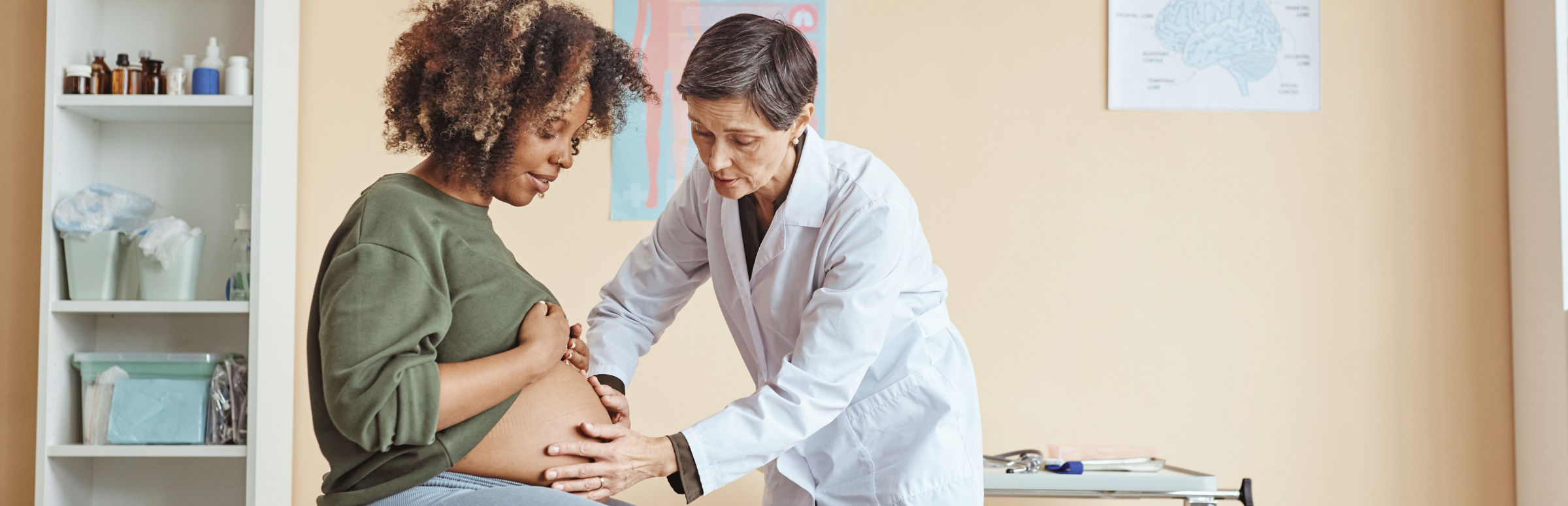 A white female doctor examines a Black pregnant woman in an exam room.