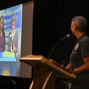 A woman at a podium watches a projected video with a smile